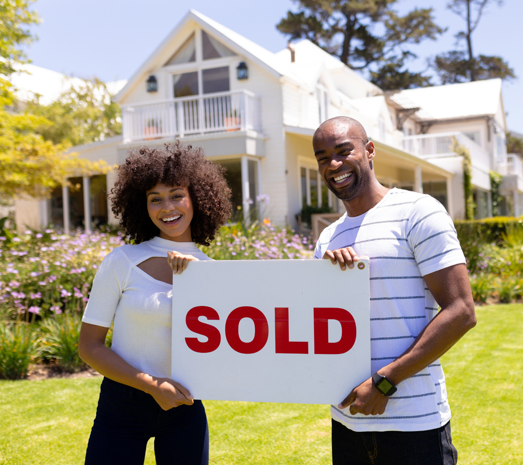 Front view of a mixed race couple standing in the garden, smiling to camera and holding sign with the word sold written on it
